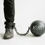 a person's lower leg and foot, shackled to a large, dark gray ball with the word ASNEF